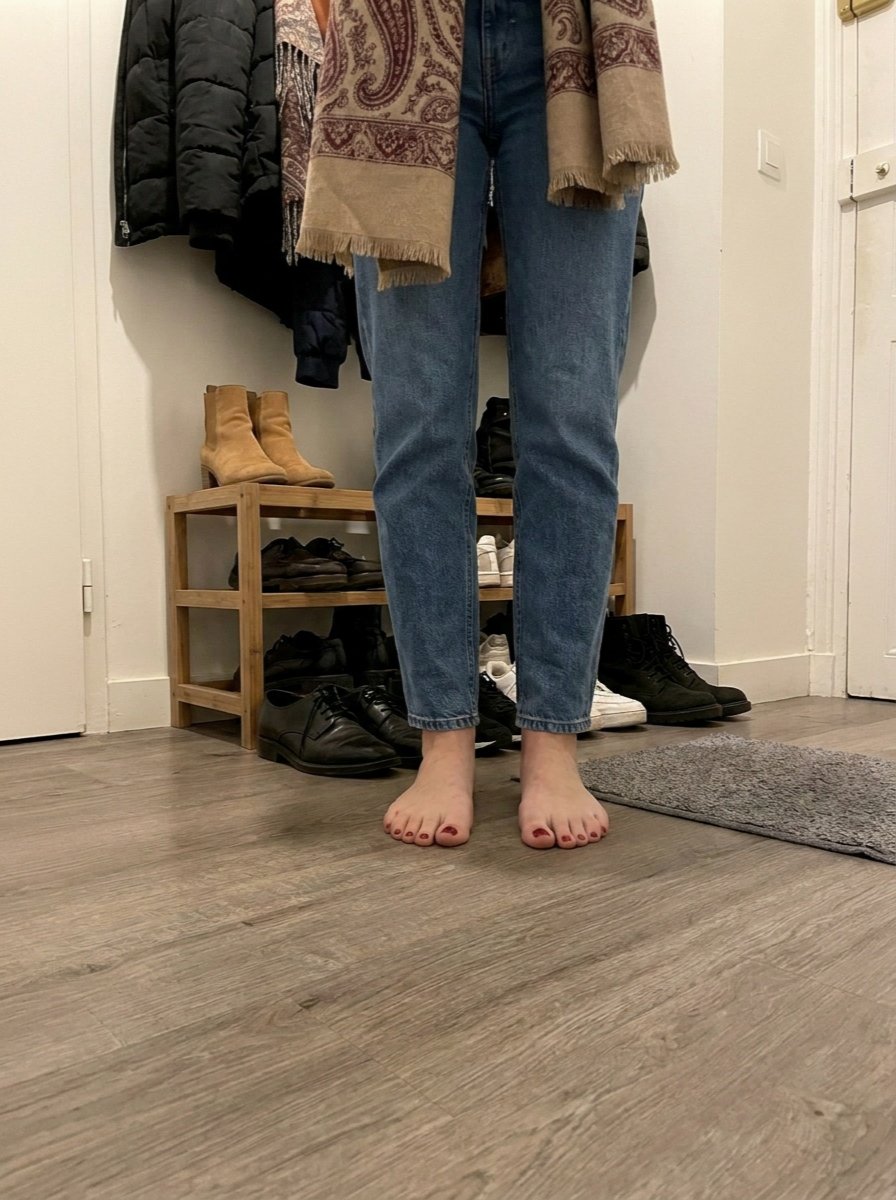 Close view of Léna’s blue jeans and bare feet with burgundy toenails as she stands by the hallway shoe rack and grey mat.