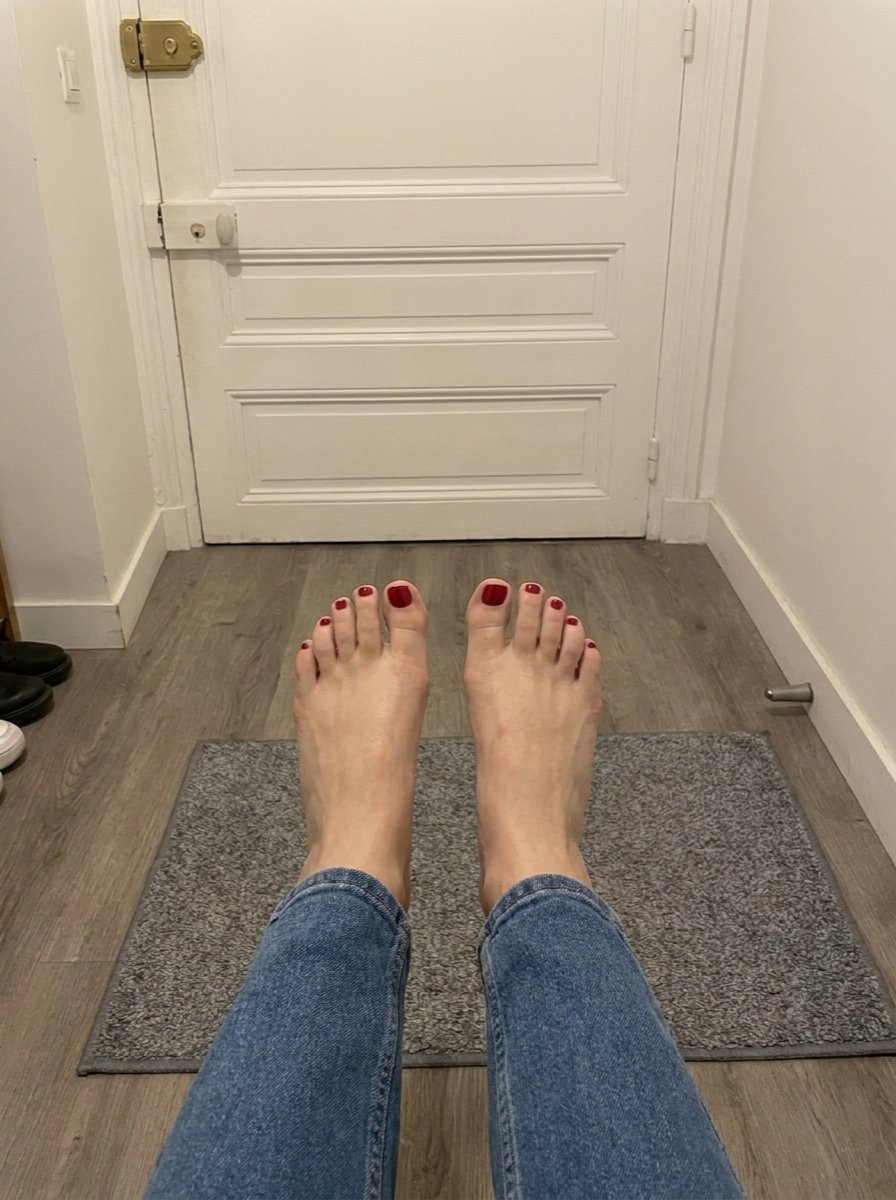 Looking down at blue-jeaned legs and bare feet with burgundy toenails, facing the grey mat and white hallway door.