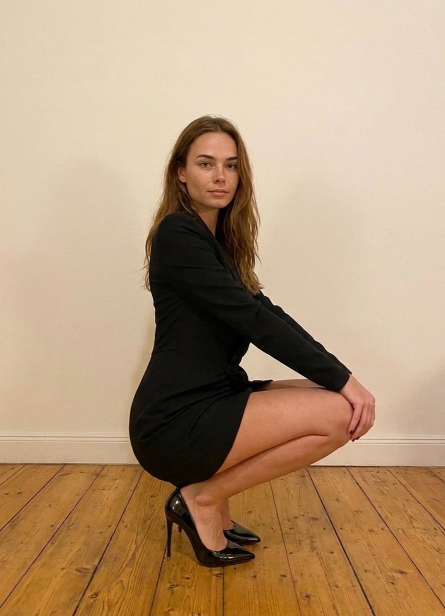 Léna crouching in profile with her hands on her knees, wearing a black blazer dress and pointed black patent pumps against a cream wall.