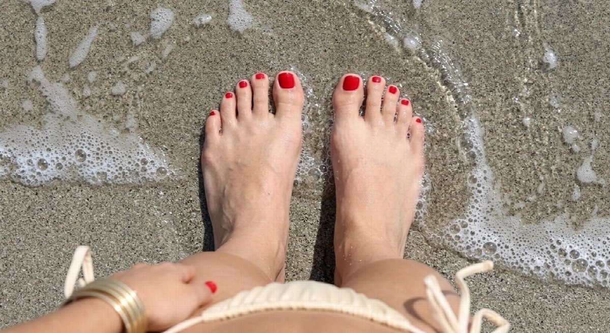 Overhead view of Lena’s bare feet with red toenails in sea foam, with her cream bikini and gold bracelets partly visible.
