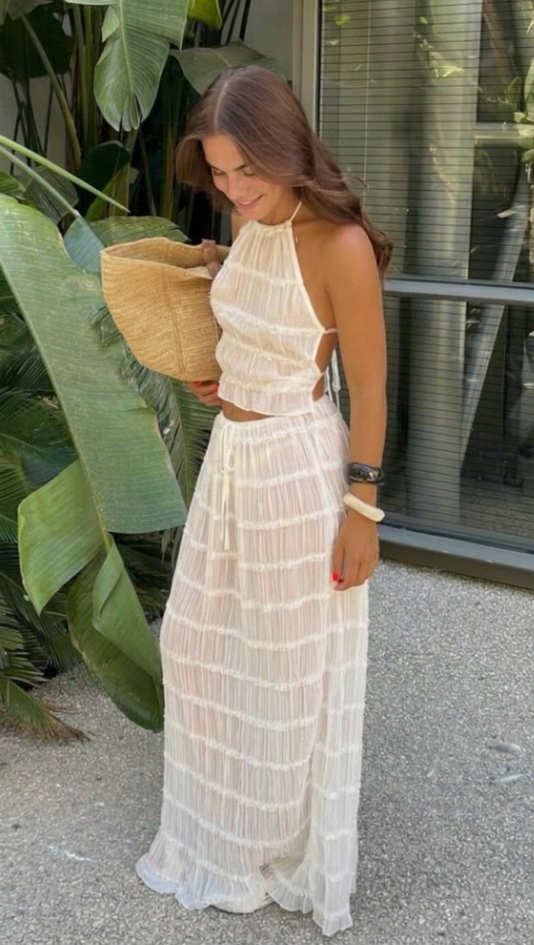 Lena standing with her gaze lowered, wearing a white crinkled halter top and long skirt, straw bag under her arm beside leaves and a window.
