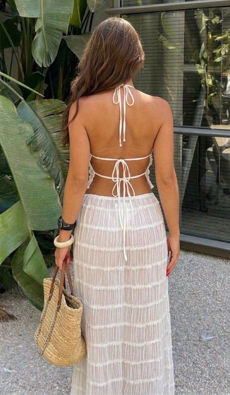 Lena standing with her back to the camera, wearing a white tie-back top and sheer long skirt, holding a straw bag beside a window.