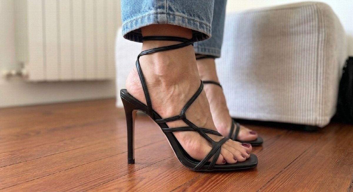 Side close-up of Lena’s feet in black stiletto sandals on the wooden floor beneath jeans, with the striped ottoman in the background.