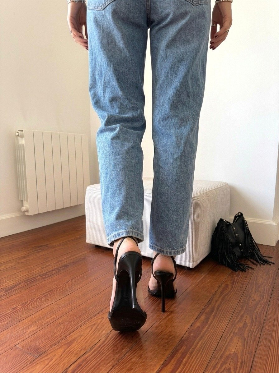 Lena is framed from behind in jeans, one foot raised in a black stiletto sandal on the wooden floor, facing the striped ottoman.