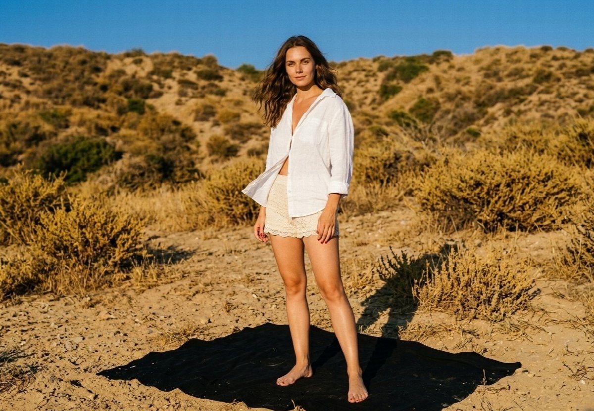 Lena stands barefoot on a black blanket, wearing an open white shirt and cream lace shorts, surrounded by sand and golden hills.