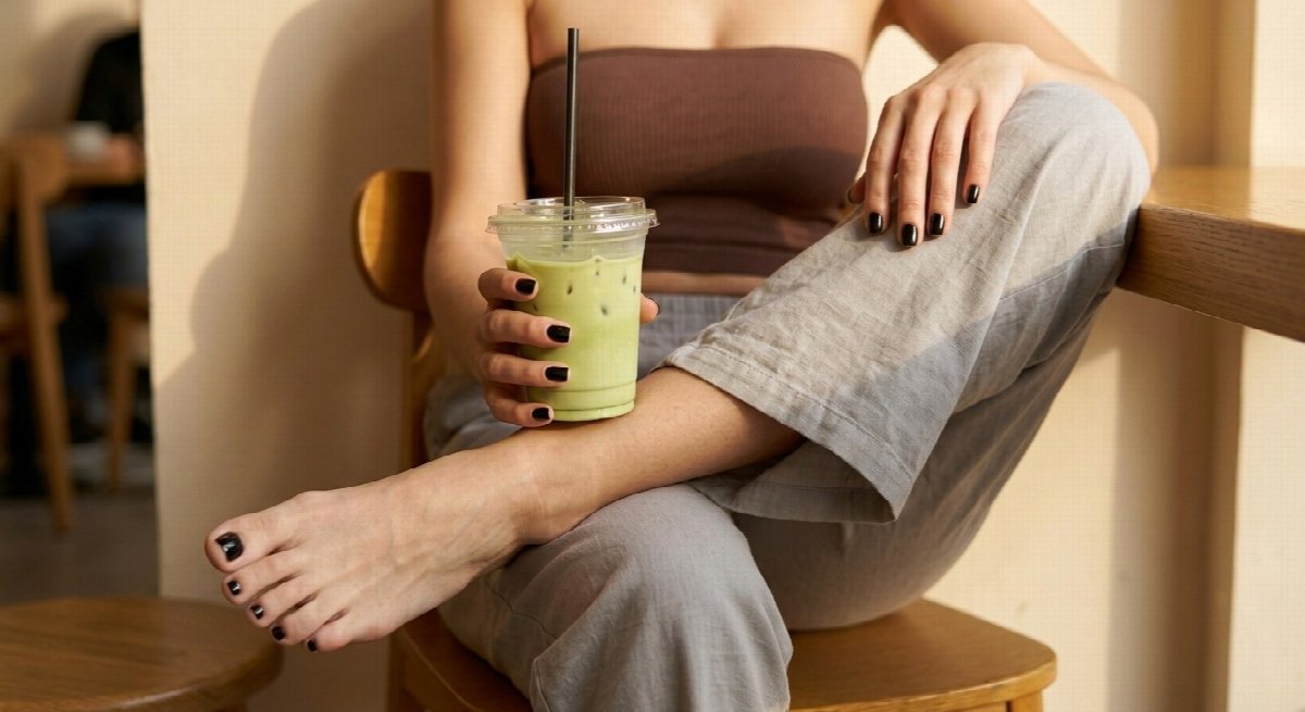 Lena is framed below the face in a brown bandeau and grey trousers, a bare foot with black polish crossed over her leg, holding matcha.