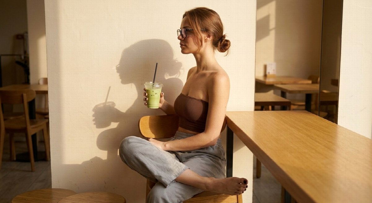 Lena sits in profile wearing black glasses, a brown bandeau and grey trousers, legs crossed and one foot bare, holding matcha beside her wall shadow.