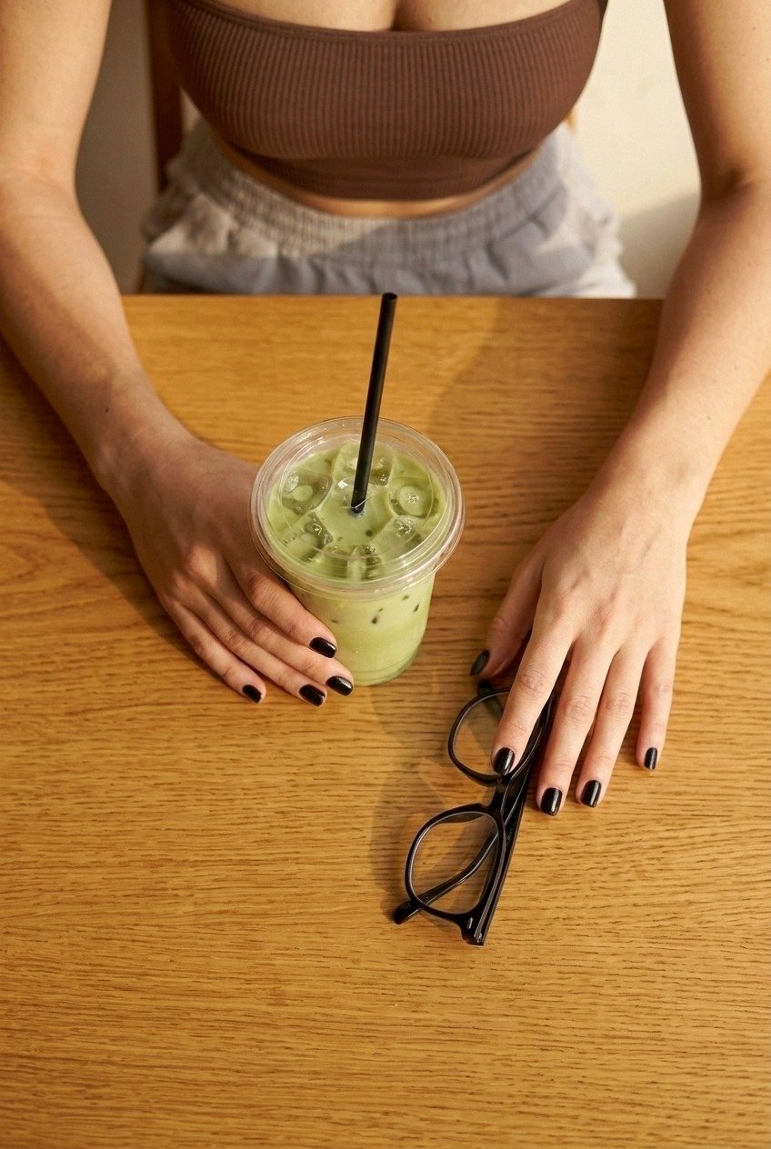 Overhead view of Lena’s hands with black nail polish, matcha and glasses on a wooden table, with her brown bandeau and grey trousers visible.