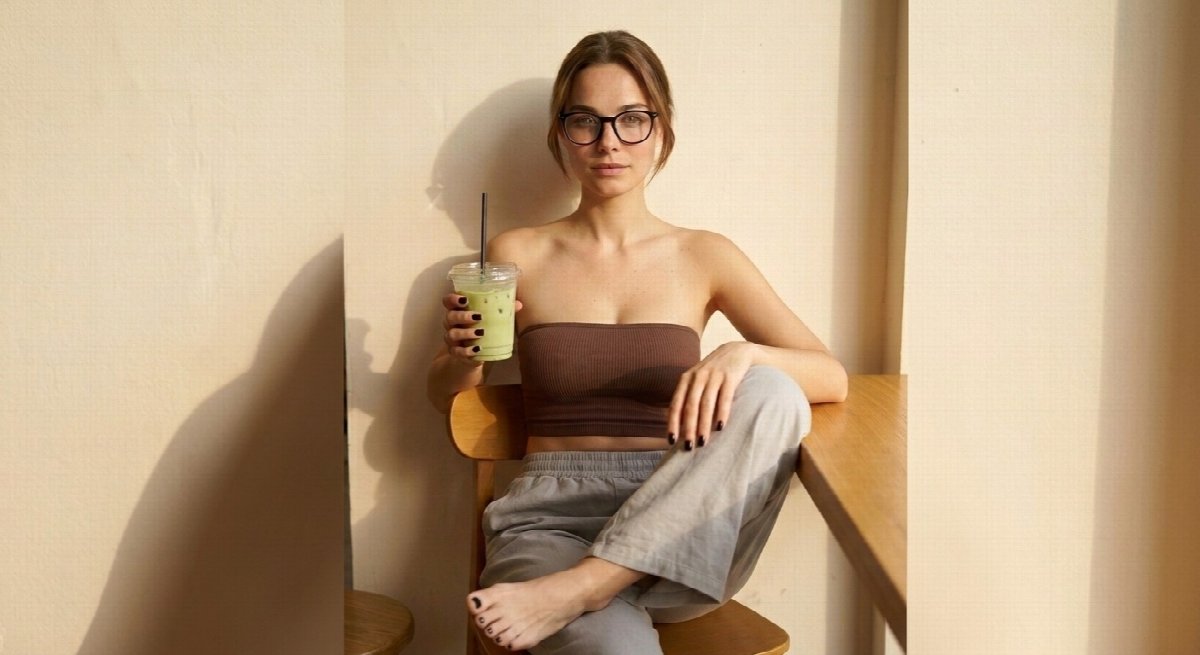 Lena sits on a chair in a brown bandeau, grey trousers and black glasses, knee raised and bare foot crossed, holding matcha between cream walls.