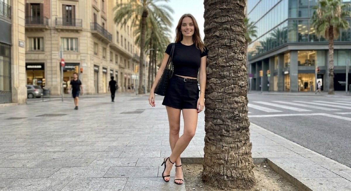 Lena stands against a palm trunk with crossed ankles, wearing a black tee, shorts and heeled sandals, with a tote over her shoulder.