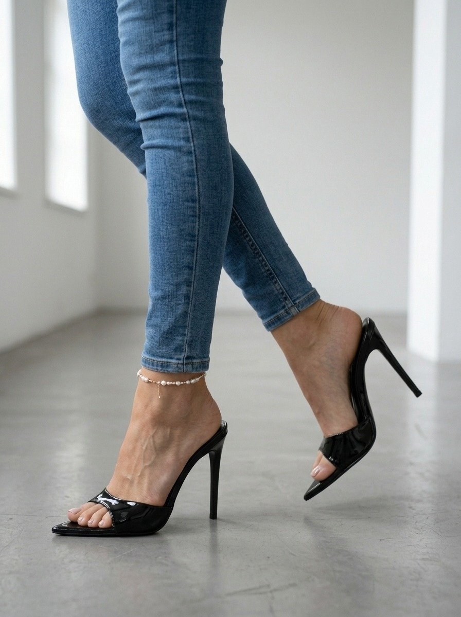 Lena's legs in skinny jeans are framed mid-step, with one black mule raised and a pearl anklet visible above the concrete floor.