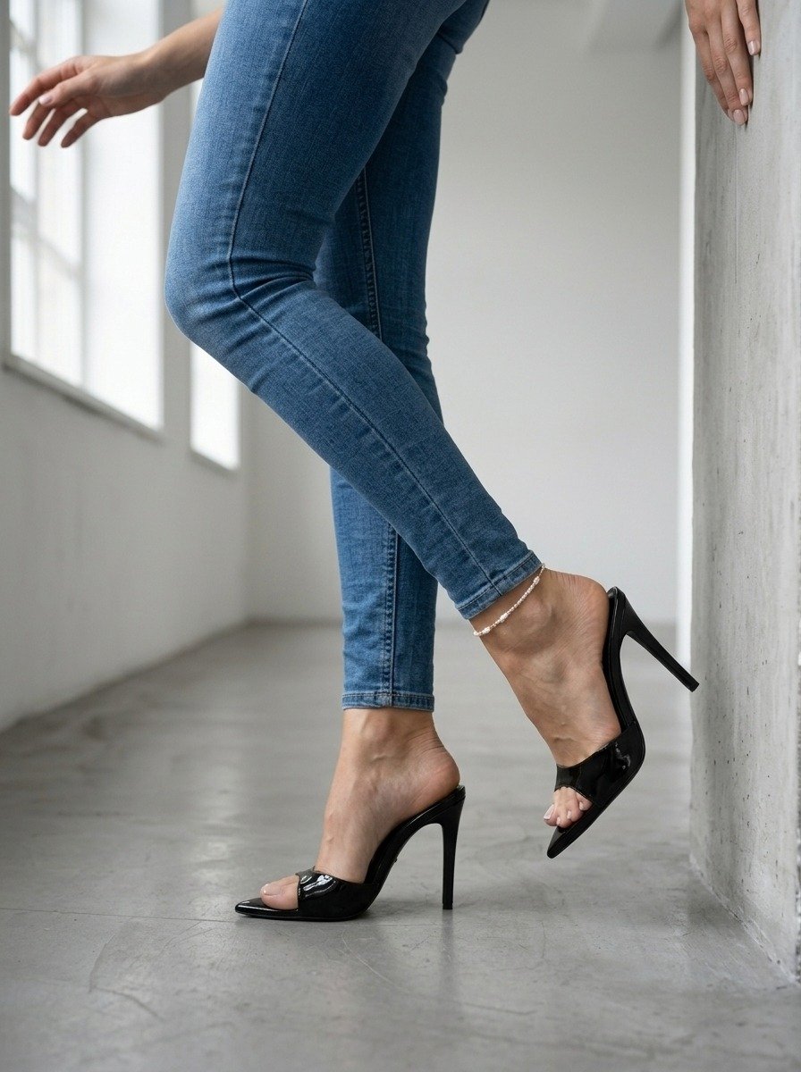 Lena is framed from waist to feet beside a wall, legs crossed in skinny jeans and black patent mules, one toe near the concrete.