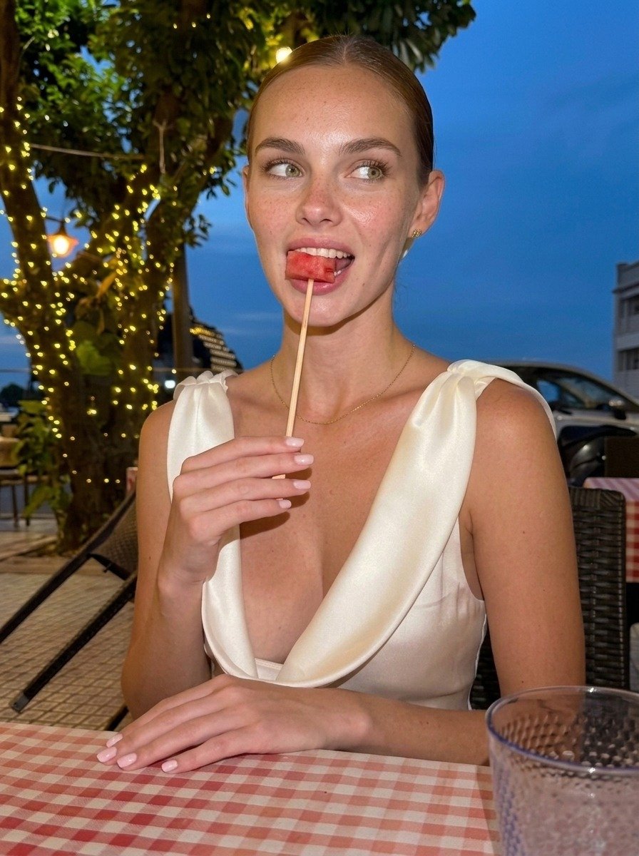 Lena in a white satin dress looks sideways, watermelon by her open mouth and one hand resting on the red and white tablecloth.