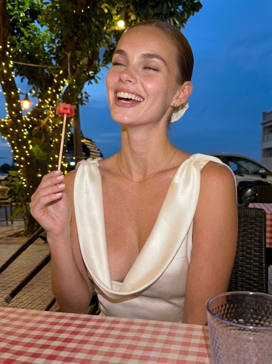 Lena in a white satin dress laughs with nearly closed eyes and her face slightly raised, holding watermelon beside the terrace lights.