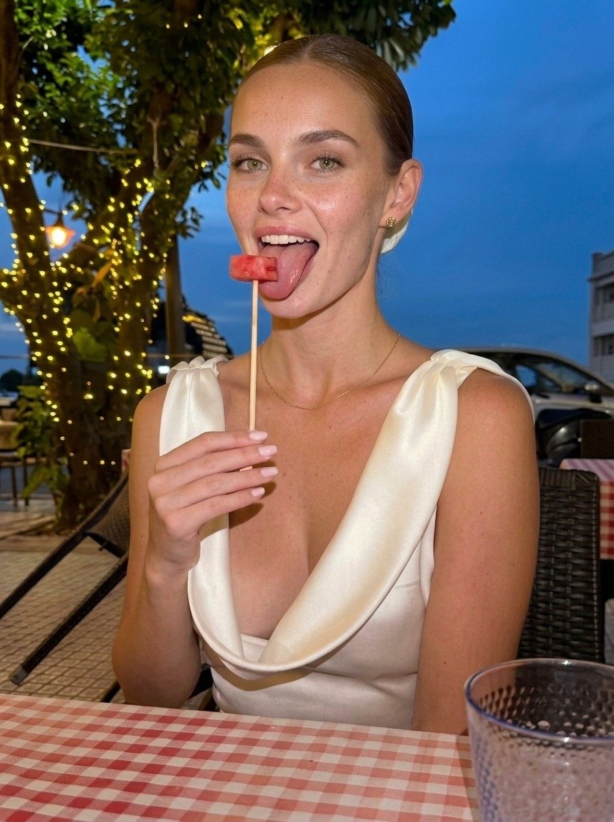 Lena in a white satin dress sticks out her tongue beside watermelon on an upright skewer, seated beneath the terrace string lights.