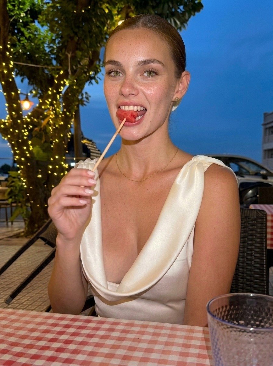 Lena wears a white dress, holding watermelon against her teeth on a diagonal skewer while seated behind the red and white gingham tablecloth.