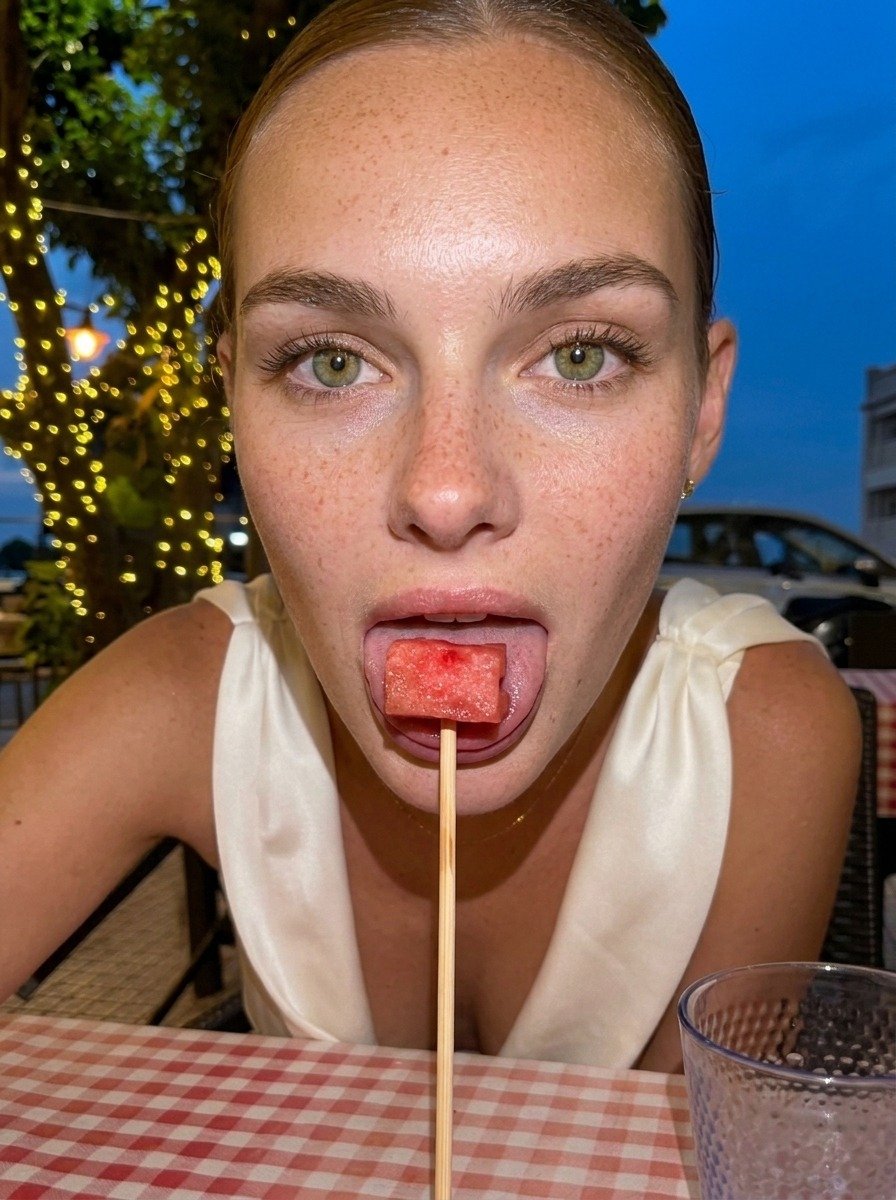 Lena leans forward in close-up, watermelon against her tongue on a vertical skewer, her white dress visible above the gingham tablecloth.