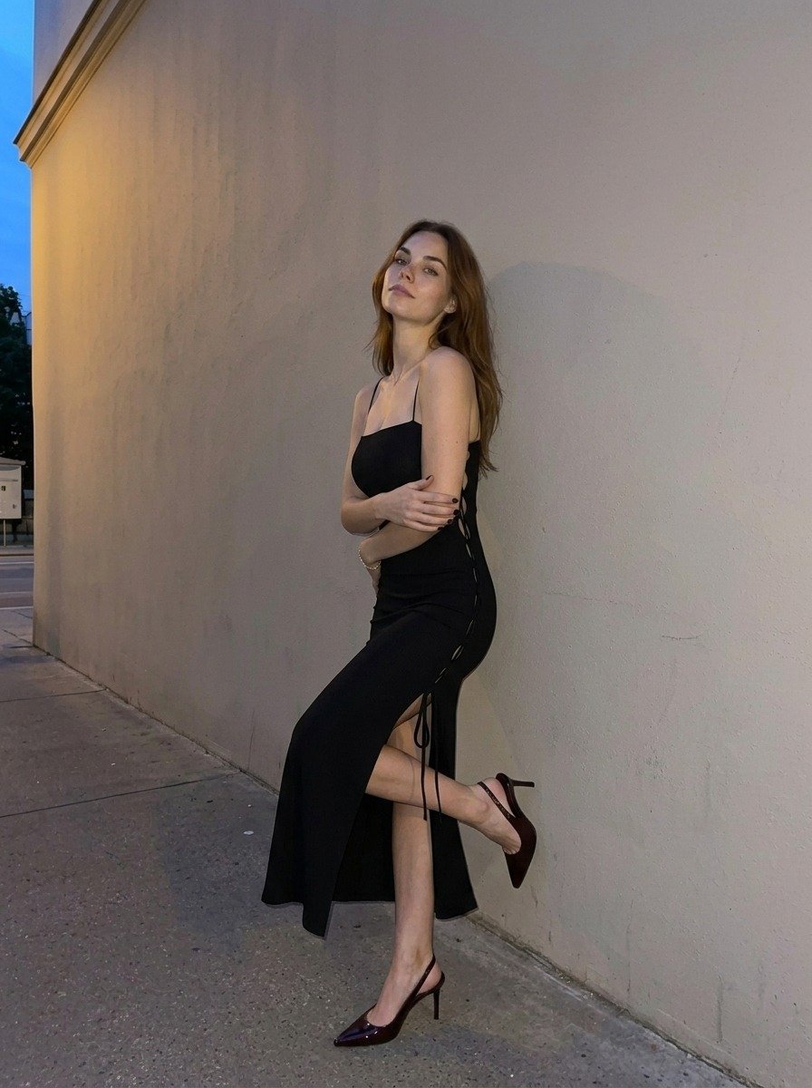 Lena leans against a beige wall with folded arms and one bent leg, wearing a black slit dress and slingback heels.