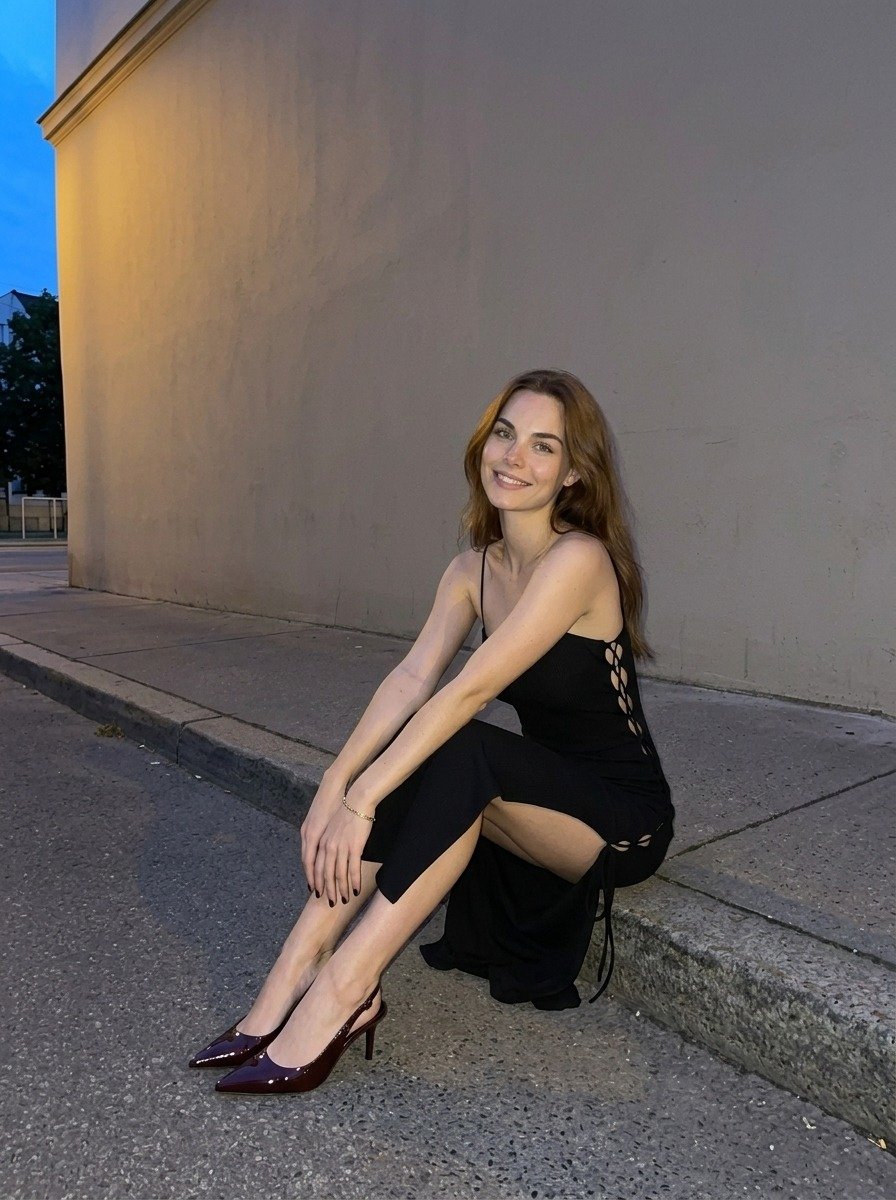 Lena sits on the curb with her hands resting on her legs, wearing a black slit dress and burgundy heels beside a beige wall.